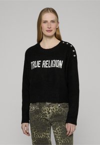 Schwarzer, gerippter Strickpullover mit der Aufschrift "TRUE RELIGION" in Weiß. Mit silbernen Knöpfen an der Schulter, kombiniert mit grünen Jeans im Leopardenmuster.