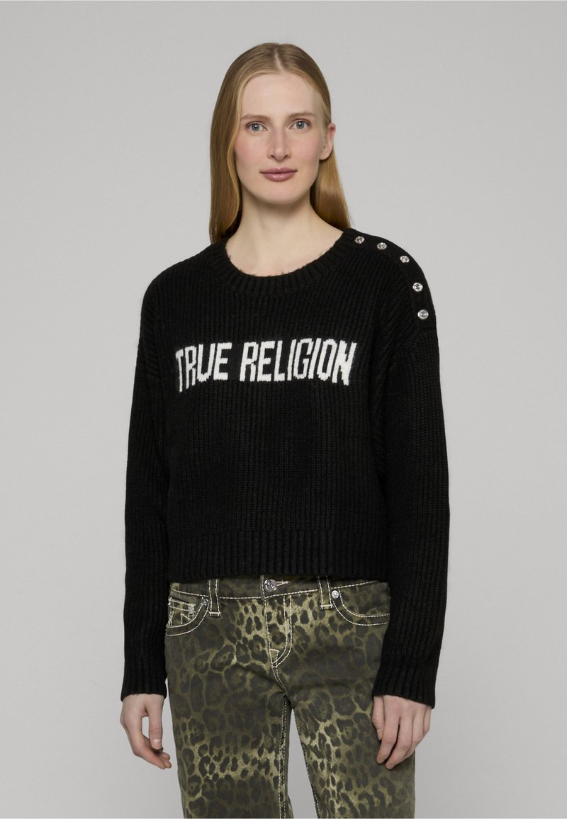 Schwarzer, gerippter Strickpullover mit der Aufschrift "TRUE RELIGION" in Weiß. Mit silbernen Knöpfen an der Schulter, kombiniert mit grünen Jeans im Leopardenmuster.