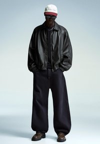 Giubbotto bomber nero in pelle con polsini a costine, pantaloni oversize neri e scarpe marroni con motivo. Cappellino da baseball bianco e rosso con logo.