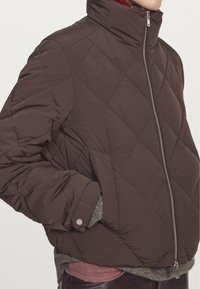 Braune, gesteppte Pufferjacke mit hohem Kragen und Reißverschluss, ausgestattet mit seitlichen Taschen und gerippten Bündchen, darunter ein grauer Strickpullover.