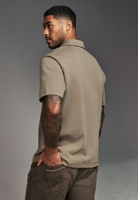 Beige poloshirt met korte mouwen en een kraag, met een gladde textuur. Het model heeft tatoeages en draagt een paarse broek met zakken.