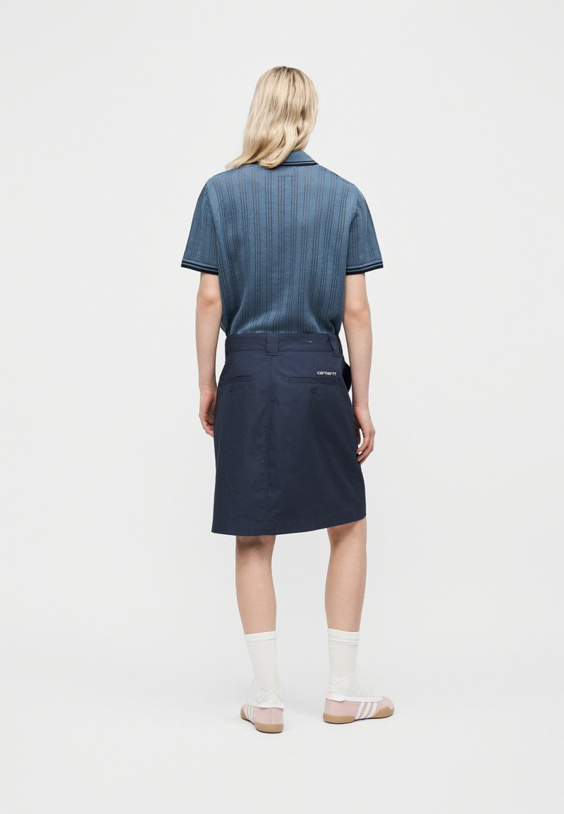 Persona con capelli biondi che indossa una maglietta a maniche corte blu in maglia, una gonna blu scuro al ginocchio con tasche, calzini bianchi e sandali beige, in piedi di spalle.