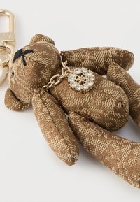 Brązowy pluszowy breloczek w kształcie misia wykonany z teksturowanej tkaniny, z haftowanymi detalami twarzy oraz złotym łańcuszkiem z zawieszką z kryształem.