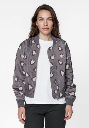 Femme aux longs cheveux bruns porte une veste bomber grise avec un motif abstrait rose clair et marron foncé par-dessus une chemise blanche et un pantalon noir.