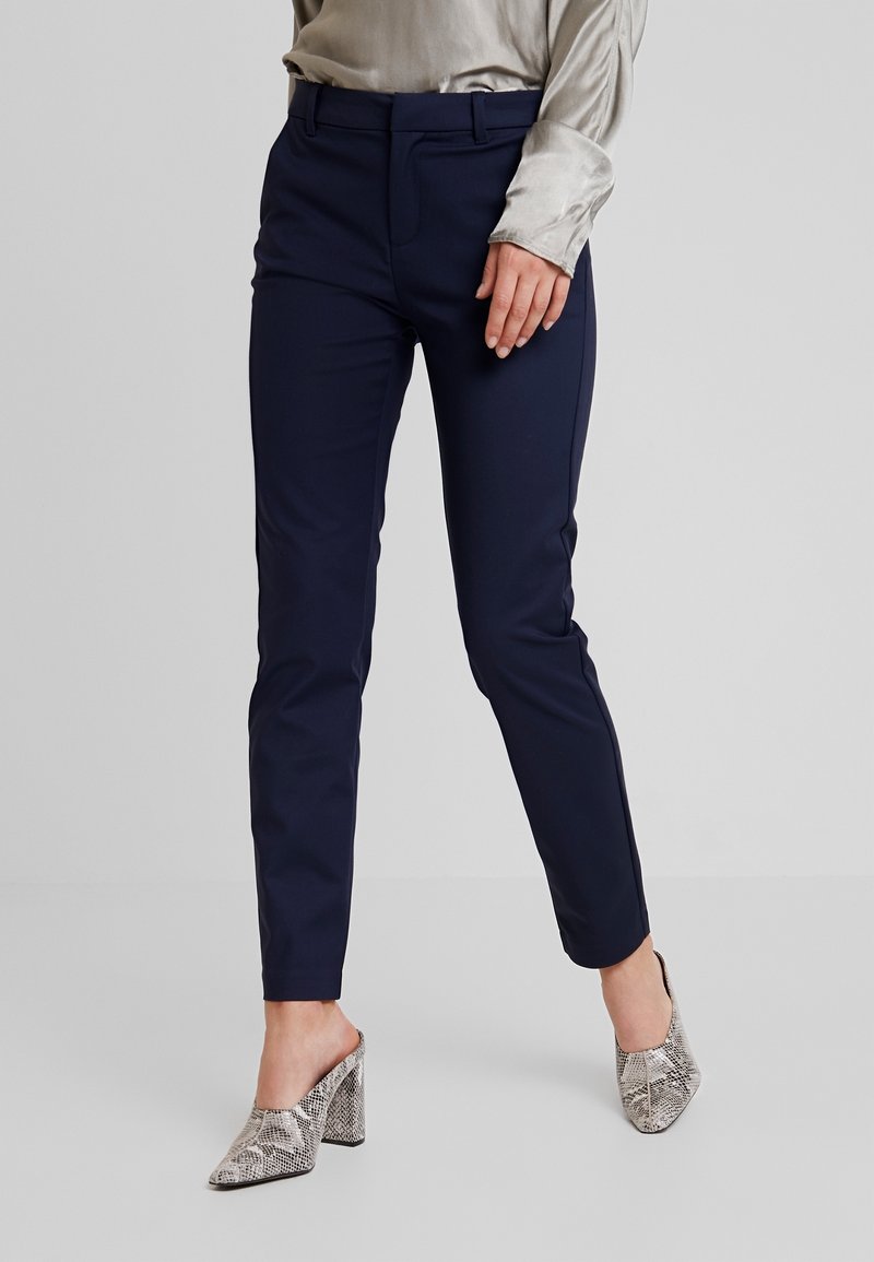 Femme portant un pantalon slim bleu marine, un chemisier en satin gris clair et des chaussures à talon bloc gris à motif peau de serpent sur un fond gris uni.