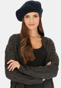 Granatowy beret z cekinami o dzianinowej strukturze, noszony z ciemnoszarym, grubym swetrem nad czarną bluzką. Modelka prezentuje neutralny wyraz twarzy.