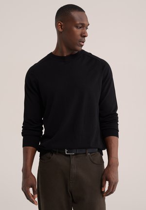 Mann trägt schwarzes Langarmshirt und dunkle Hose, steht und schaut nach links vor einem einfarbigen hellen Hintergrund.