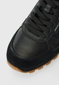 Gros plan sur l'avant d'une sneaker en cuir noir et en suédine avec des lacets noirs et une semelle texturée marron sur fond blanc.