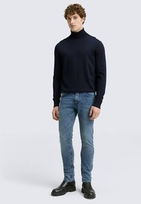 Marineblauer Rollkragenpullover aus feinem Strick, kombiniert mit hellblauen Slim-Fit-Jeans und schwarzen Stiefeletten. Das Modell steht vor einem neutralen Hintergrund.