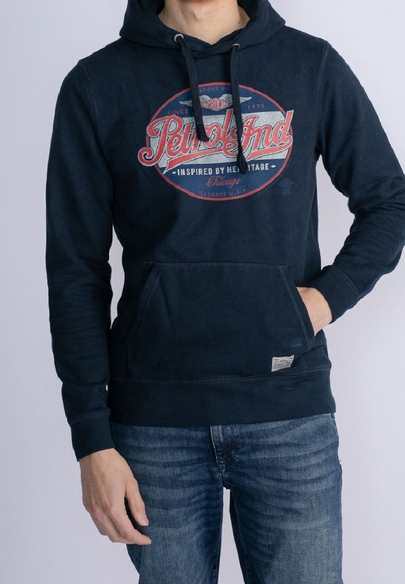 Homme portant un sweat à capuche bleu marine avec le logo rouge et blanc "Petrolind" et un jean bleu, une main dans la poche du sweat, sur fond clair.