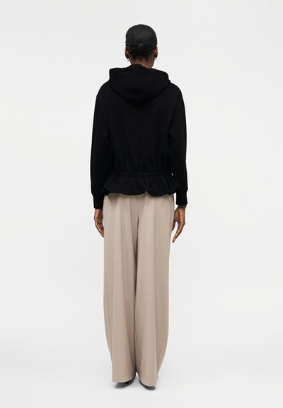 Γυναίκα με μαλλιά σε κότσο φοράει μαύρο μπουφάν με κουκούλα και βολάν στο τελείωμα και άνετο μπεζ παντελόνι με φαρδιά μπατζάκια, εμφανίζεται από πίσω.