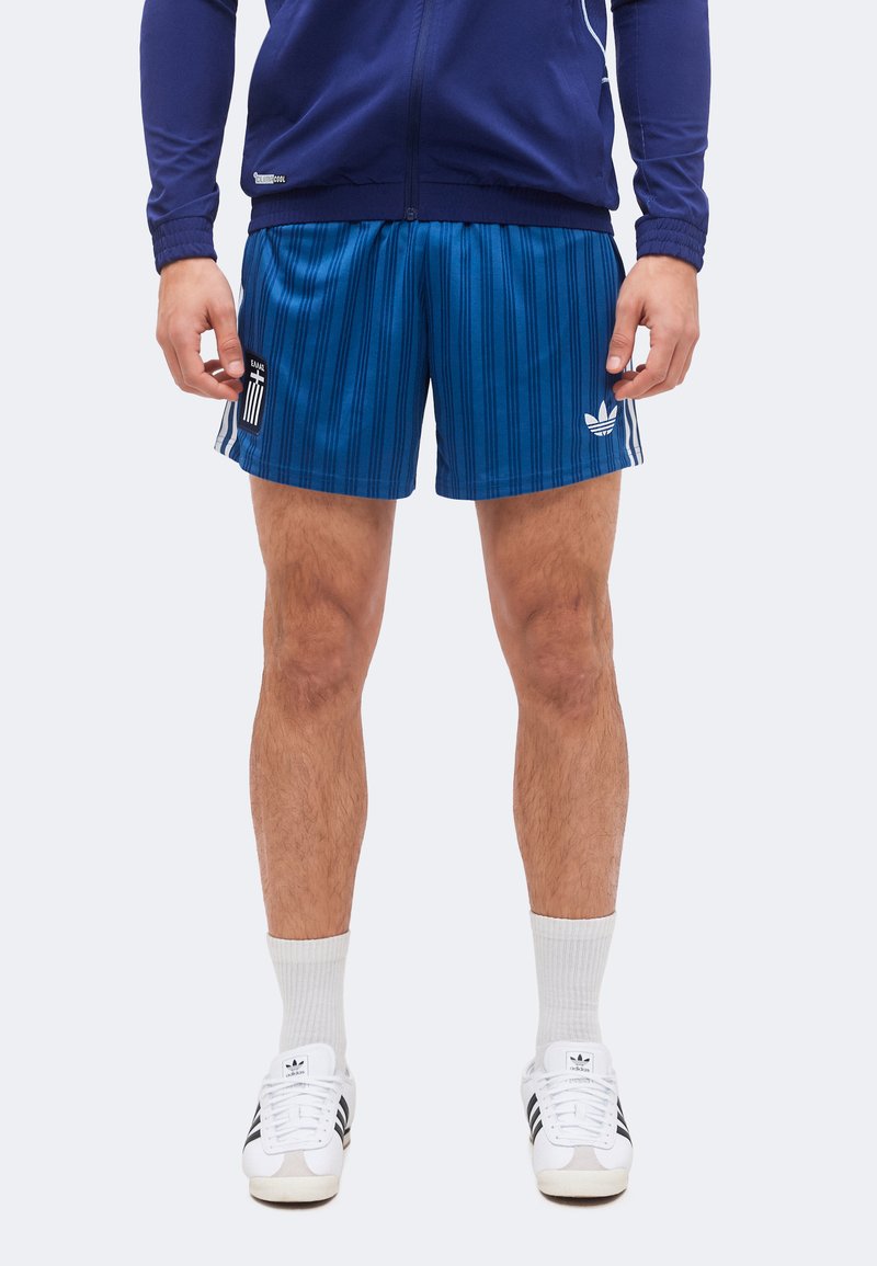 Man draagt een donkerblauwe jas met rits, blauwe gestreepte sportshort met logo's, witte Adidas-sokken en witte Adidas-sneakers, rechtopstaand.