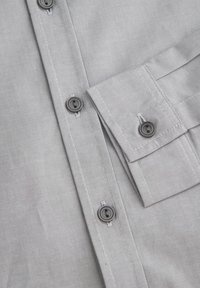 Szara koszula na guziki wykonana z delikatnie teksturowanej tkaniny, z dwoma guzikami z przodu oraz kieszenią z klapką ozdobioną kontrastowym przeszyciem.