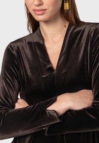 Hnedé zamatové šaty s hlbokým výstrihom do V a dlhými rukávmi s rozparkom. Modelka má na sebe veľké zlaté obdĺžnikové náušnice.