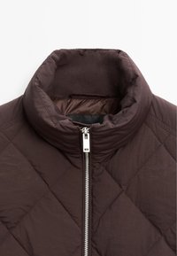 Braune gesteppte Jacke mit hohem Kragen und silbernem Reißverschluss. Sie hat eine glatte Textur und ein Rautenmuster auf dem Stoff.