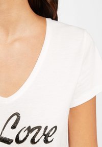 Maglietta bianca in cotone con scollatura a V, caratterizzata da un design grafico in paillettes nere con la scritta "Love" sul davanti e maniche corte.