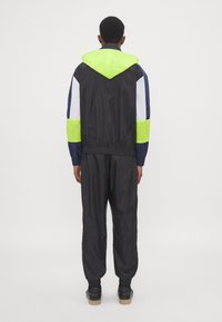 Black windbreaker with neon green accents and mesh panels, featuring a hood and elastic cuffs, paired with matching black trousers.