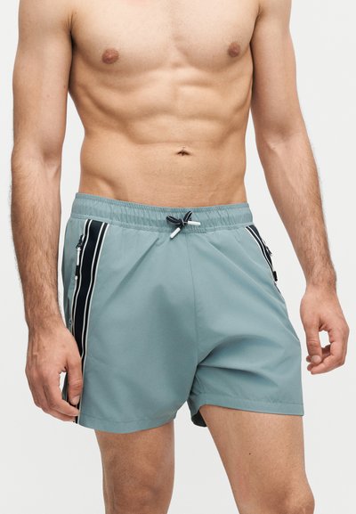 Ανδρικός κορμός με ανοιχτό μπλε μαγιό βερμούδα με μαύρες και λευκές πλαϊνές ρίγες και κορδόνι στη μέση, στέκεται μπροστά σε λευκό φόντο.