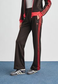 Μαύρο παντελόνι φόρμας με κόκκινες ρίγες στο πλάι, με τσέπη με φερμουάρ και λογότυπο Adidas. Συνδυάζεται με γκρι αθλητικά παπούτσια με λευκές λεπτομέρειες.