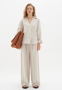 Top in lino con scollo a V e maniche a costine di colore beige chiaro, abbinato a pantaloni beige a gamba larga. La modella porta una grande borsa marrone.