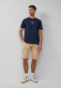 Marine-kleurige katoenen t-shirt met witte tekst, gecombineerd met beige cargoshorts met grote zijzakken en beige sneakers met crème accenten.