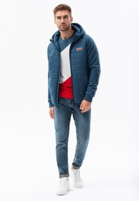 Giacca blu imbottita con cappuccio, chiusura con zip e polsini elastici. Abbinata a un maglione a collo rotondo color block e jeans slim-fit, completata da sneakers bianche.