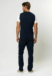 Temno modra pletena brezrokavnik nad belim majico, nosi se s temnimi denim hlačami in svetlo sivo obutvijo, prikazan od zadaj.