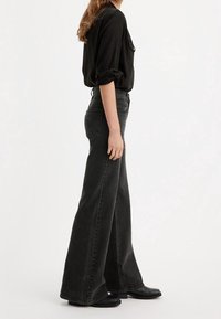 Pantalons en denim noir évasés avec une taille haute, dotés de passants de ceinture et de poches arrière, portés avec une chemise noire à manches longues et des bottes noires.