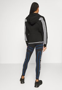 Sudadera negra con rayas blancas en las mangas, combinada conleggings negros con acentos azules y texto blanco. Se puede ver la textura de la tela.