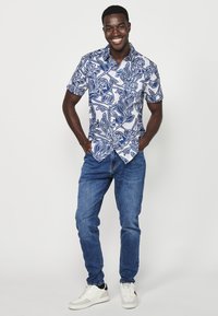 Camicia a maniche corte con bottoni, motivo floreale blu e bianco, abbinata a jeans blu e sneakers bianche, in piedi contro uno sfondo bianco.