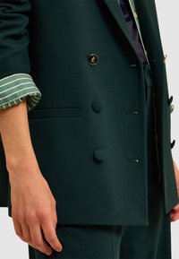 Blazer verde scuro con una trama liscia, design doppiopetto, caratterizzato da bottoni rotondi e una tasca discreta. Maniche a righe visibili.