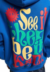 Sudadera con capucha azul con texto llamativo y divertido en amarillo, verde, rojo y azul, con un gráfico de sol y las letras "KK" en un círculo.