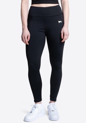 Osoba nosiaca čierne legíny s vysokým pásom s malým bielym levím logom na bokoch a bielou športovou obuvou, stojaca pred jednoliatym pozadím.