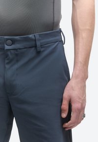 Nahaufnahme einer Person, die marineblaue Hosen mit Knopfverschluss, Gürtelschlaufen und einem grauen, eng anliegenden Oberteil trägt, wobei die rechte Hand mit einem Ring gezeigt wird.