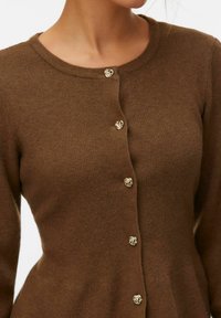 Cardigan en maille marron avec encolure ronde et six boutons décoratifs dorés en forme de nœud sur le devant, porté par une personne visible du cou à la taille.