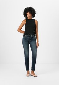 Zwarte geribde tanktop gecombineerd met donkerblauwe slim-fit jeans en lichtroze flats, tegen een egaal witte achtergrond.