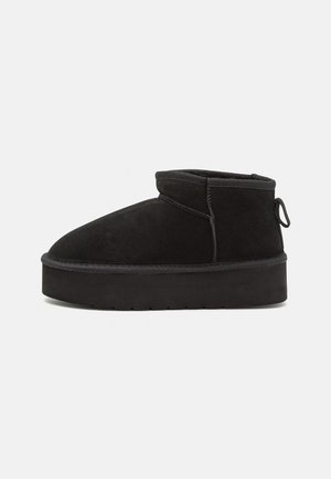 Black suede ankle boot with a rounded toe, featuring a raised sole and a side loop for easy wear. The design is simple with minimal accents.
