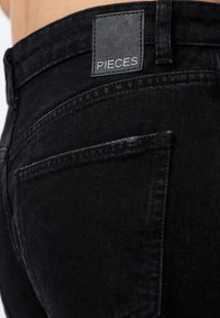 Nærbilde av baklommen på svarte jeans i denim med et sort skinnmerke på linningen med teksten "PIECES".