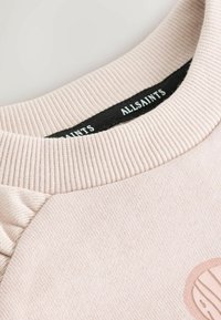 Világos rózsaszín pulóver bordázott nyakkivágással és texturált anyaggal. Elöl enyhén sötétebb árnyalatú kör alakú logóval díszített.