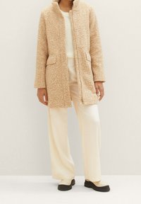 Beige fleecejacka med en texturerad yta, dragkedjestängning, två framfickor och ståkrage, bärs över en ljus tröja och krämfärgade byxor.