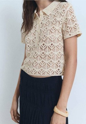 Kvinde iført en beige kortærmet hæklet top med krave og knapper, kombineret med et sort tekstureret højhalset skørt og et beige armbånd.