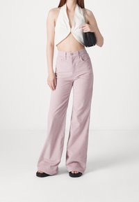 Femme portant un crop top blanc à col halter, un pantalon rose large taille haute, des sandales noires et tenant un petit sac à épaule noir.