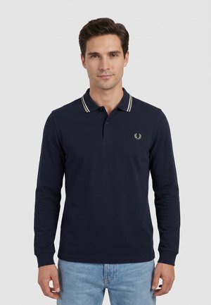 Uomo che indossa una polo blu navy a maniche lunghe con colletto a righe bianche e jeans azzurri, in piedi davanti a uno sfondo semplice.