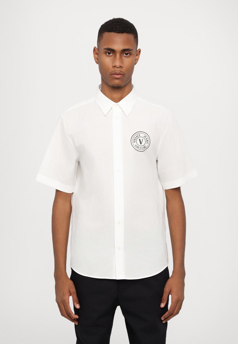 Chemise blanche à manches courtes avec boutons, fabriquée en tissu léger, dotée d'un patch logo noir rond sur la poitrine gauche et d'un col classique.