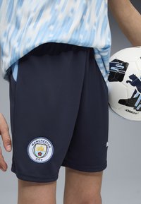 Jūras spēku sporta šorti ar teksturētu audumu, ar Mančestras pilsētas logo uz apakšmales. Kombinēti ar gaiši zilu svītrotu krekla un futbolu.