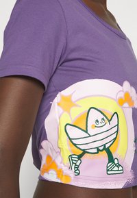Fioletowy krótki top z krótkim rękawem z kolorową, kreskówkową grafiką przedstawiającą uśmiechniętą postać na pastelowym gradientowym tle.