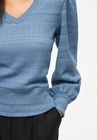 Blauw gebreid topje met een V-hals, structuurstof met horizontale strepen en pofmouwen. Gecombineerd met zwarte broeken.