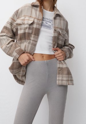 Woman wearing beige and brown plaid jacket, white cropped graphic tee, and high-waisted ribbed grey leggings, holding jacket open at waist.
