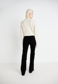 Přesně veliký krémový svetr, měkká pletenina. Černé zvonové džíny, hladká tkanina. Modelka viděna zezadu, minimalistické prostředí.