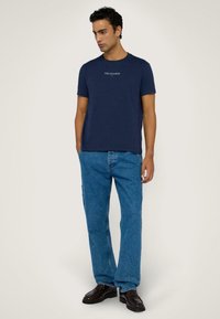 Camiseta de algodón de color azul marino con cuello redondo y logo blanco "TRUSSARDI". Combinada con jeans de corte relajado en azul y zapatos negros.